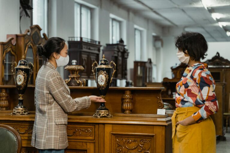 Two women in face masks interacting in an antique store with vintage furniture.
